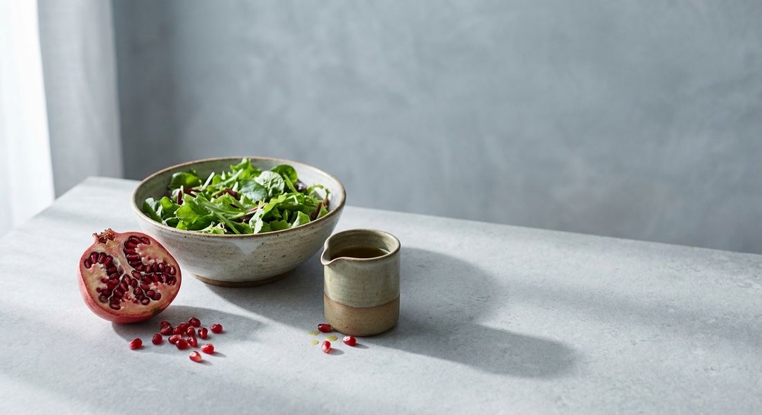 A halved pomegranate with its seeds spilled on a stone counter beside a salad bowl, a fruit on the Tayyibat diet yes list